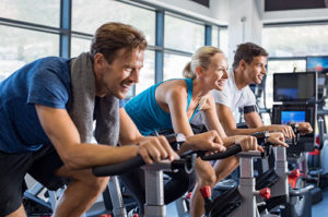 Group of smiling friends at gym exercising on stationary bike. Happy cheerful athletes training on exercise bike. Young men and woman working out at a class in the gym.